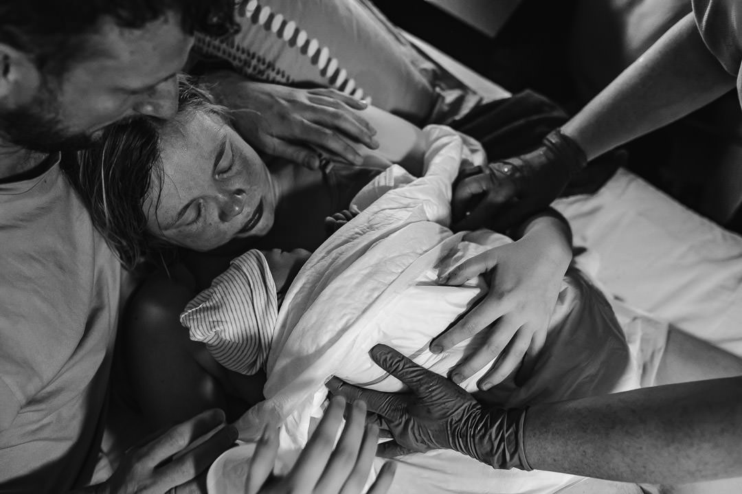 Geboorte Febe Zwart wit foto. Febe is net geboren en ligt gewikkeld in doeken bij mama op de borst. Mama kijkt gelukkig en opgelucht, papa heeft zijn arm om mama geslagen en kijkt trots naar zijn dochter. De handen van de verloskundige zijn ook in beeld om nog een extra deken over Febe heen te leggen.