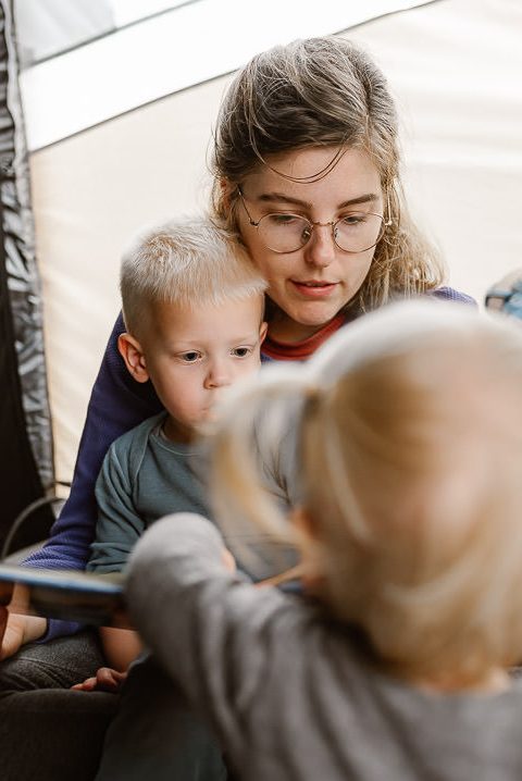 Lotte leest een boekje voor aan haar kinderen in de tent. Haar zoon zit op schoot en haar dochter staat er naast